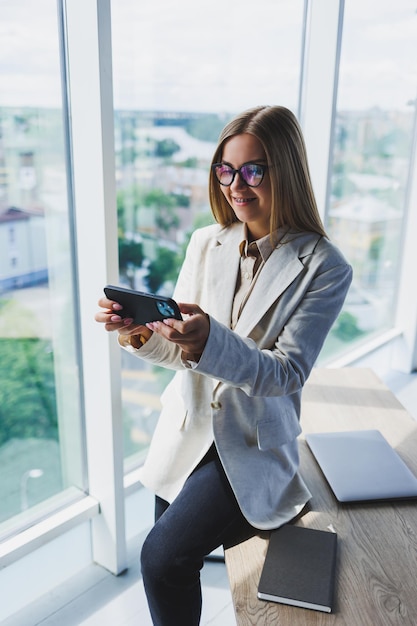 Cheerful young beautiful woman in glasses talking on a mobile phone and using a laptop with a smile while sitting at her workplace Work in the office