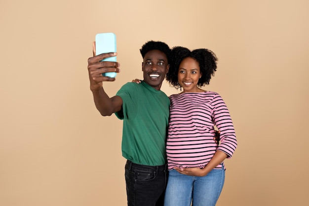 Cheerful young black man hugging lady with big belly make selfie on smartphone isolated on beige