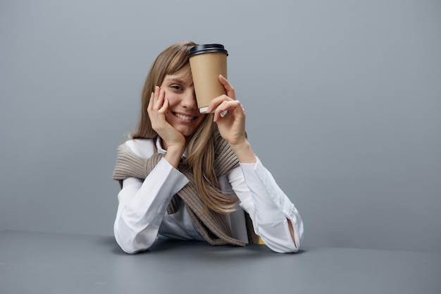 Cheerful young blonde student lady freelancer in warm sweater with takeaway coffee smile at camera sitting at workplace in gray modern home office Coffee break Lover Concept Copy space Cool offer