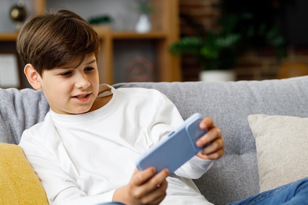 Cheerful young boy playing video game on smartphone sitting on sofa at home Cute teenager playing in car racing game on mobile phone Spending time at home leisure relax happy carefree childhood