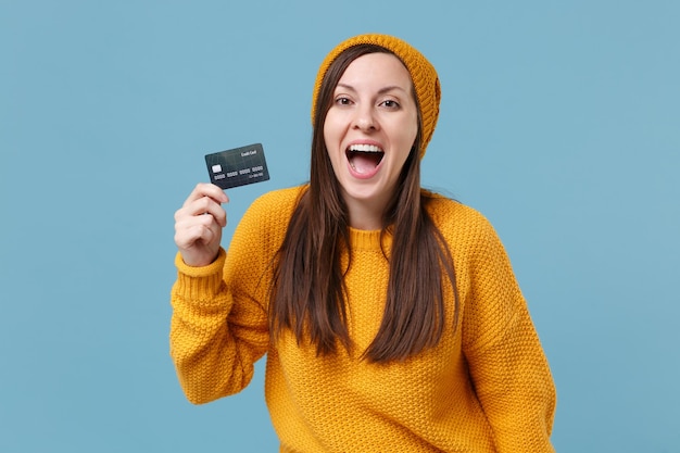 Cheerful young brunette woman girl in yellow sweater and hat posing isolated on blue background studio portrait. People sincere emotions lifestyle concept. Mock up copy space. Hold credit bank card.