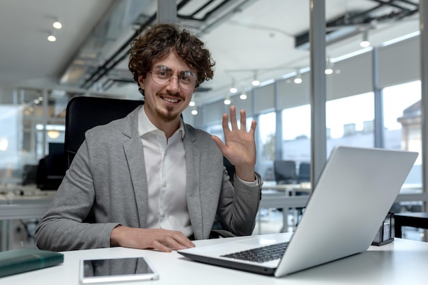 Cheerful young businessman with curly hair waving and smiling in a bright modern office setting