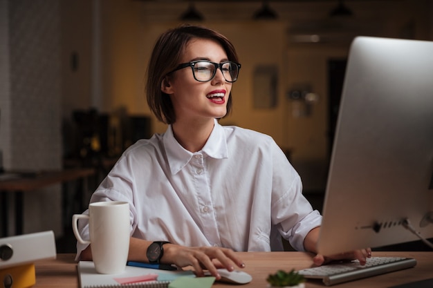 Cheerful young businesswoman in glasses using computer and smiling in office