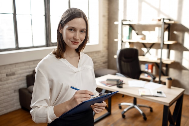 Cheerful young businesswoman stand in room and look on camera. She hold plastic folder with document and write.