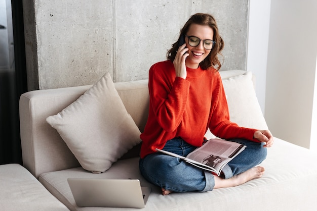 Cheerful young casually dressed woman in headphones sitting on a couch at home, studying with laptop computer, holding mobile phone