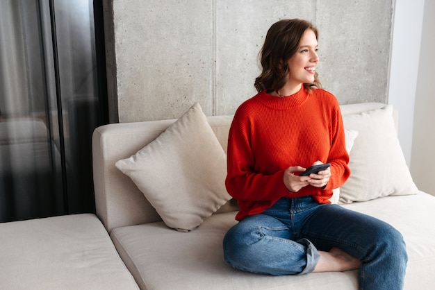 Cheerful young casually dressed woman sitting on a couch at home, holding mobile phone