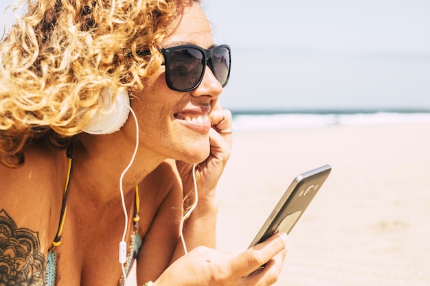 cheerful young caucasian blonde woman with sand on her faces  on the beach listening to music