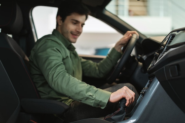 Cheerful young Caucasian guy test driving new car before purchase at automobile dealership selective focus