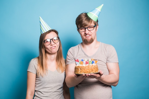 Cheerful young couple charming guy and cute girl in paper hats make foolish face and hold in their hands a cake with the inscription birthday standing on a blue wall. Concept greetings and prank