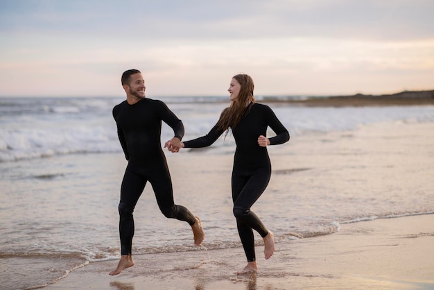 Cheerful young couple wearing wetsuits running on seashore together