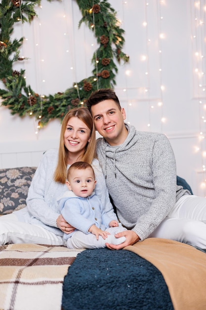 A cheerful young couple with a little son are playing on the bed near the Christmas tree New Year's interior in the bedroom Christmas tree with toys Festive family atmosphere