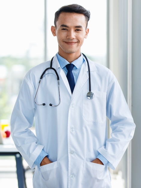 Cheerful young  and cute Asian male doctor in uniform and with stethoscope standing in clinic and pose with positive and self confident gesture.