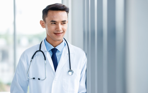 Cheerful young  and cute Asian male doctor in uniform and with stethoscope standing in clinic and pose with positive and self confident gesture.