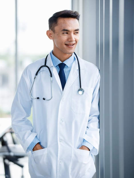 Cheerful young  and cute Asian male doctor in uniform and with stethoscope standing in clinic and pose with positive and self confident gesture.