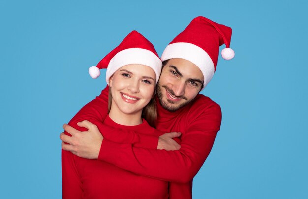 Photo cheerful young european couple in santa hats embracing and smiling at camera