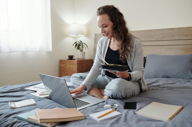 Cheerful young female entrepreneur sitting on bed in her bedroom eating tasty lunch and working on l...