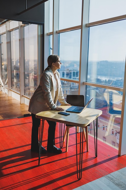 Cheerful young female freelancer in casual clothes standing at a wooden table with a netbook and writing notes while working on a project Modern young business lady in the office