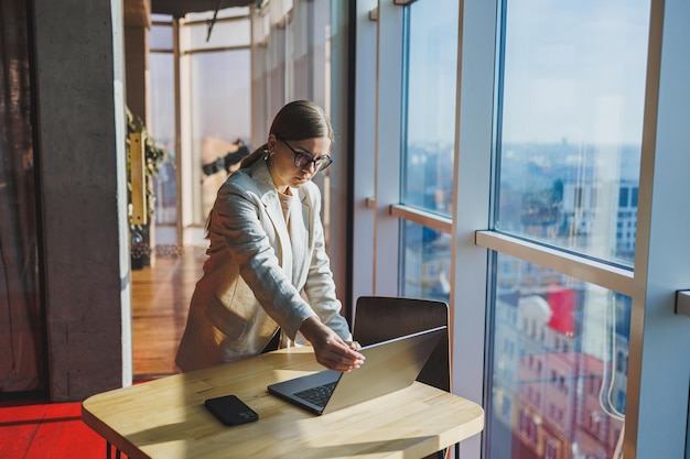 Cheerful young female freelancer in casual clothes standing at a wooden table with a netbook and writing notes while working on a project Modern young business lady in the office