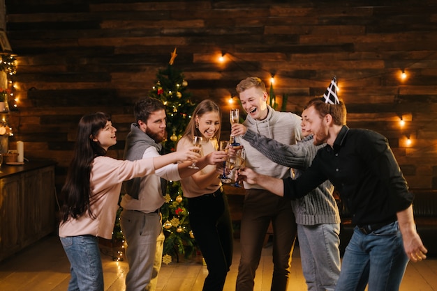 Cheerful young friends are clinking with champagne glasses and celebrating New Year. Christmas tree with garland and festive illumination in background. Friends are celebrating Christmas Eve.