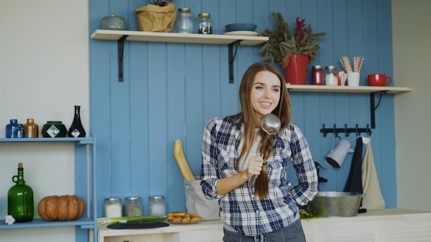 Photo cheerful young funny woman dancing and singing with ladle while having leisure time in the kitchen