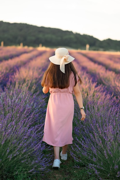 A cheerful young girl in a pink dress and a straw hat in her hands stands among lavender bushes