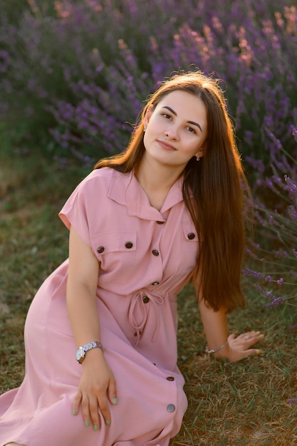 A cheerful young girl in a pink dress and a straw hat in her hands stands among lavender bushes