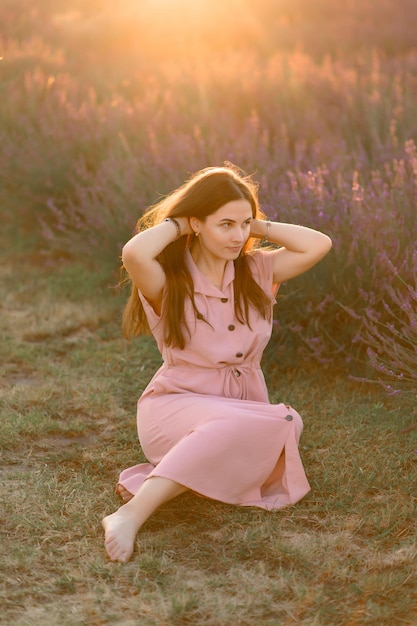 A cheerful young girl in a pink dress and a straw hat in her hands stands among lavender bushes