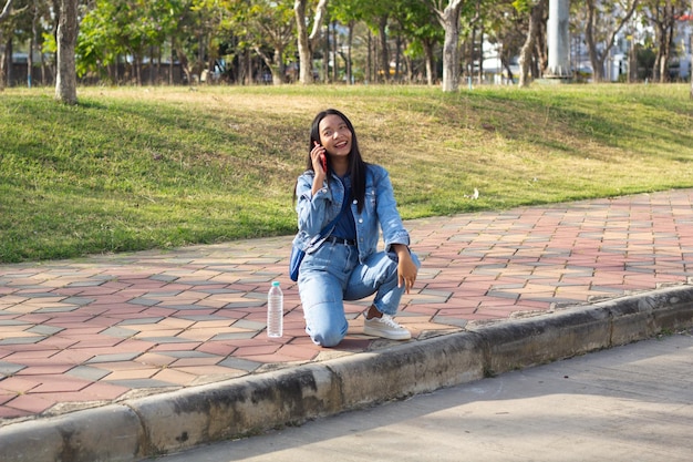 Cheerful young girl sitting at the park in city hold mobile phone with a water bottle placed on the side lifestyle concept