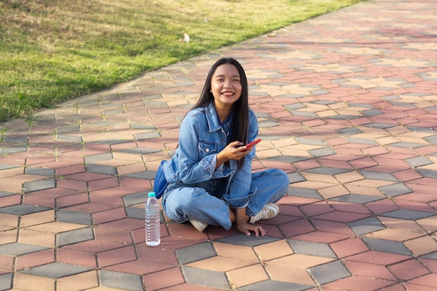 Cheerful young girl sitting at the park in city hold mobile phone with a water bottle placed on the side lifestyle concept