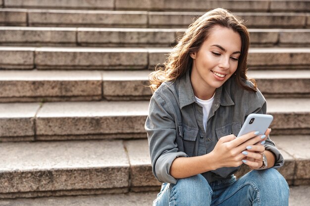 Photo cheerful young girl sitting on stairs outdoors, using mobile phone