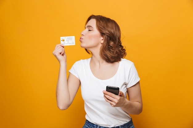 Cheerful young girl wearing t-shirt standing isolated over yellow wall, using mobile phone, showing plastic credit card