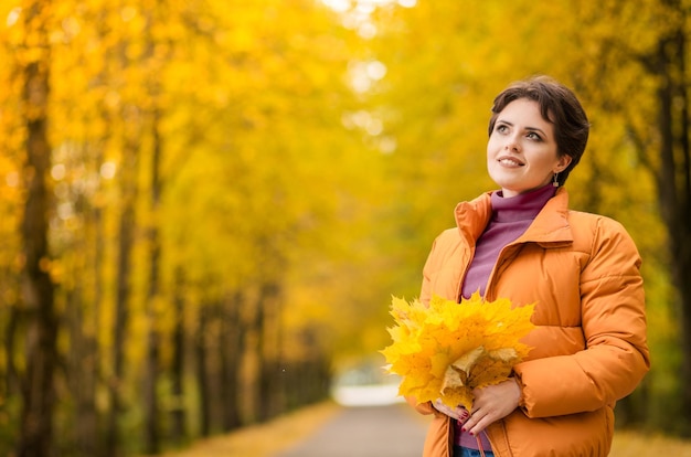 Cheerful young girl with short brown hair wearing autumn yellow jacket walking at the park Autumn walk Woman portrait Girl is playing with leaf and smiling Woman enjoying fall nature