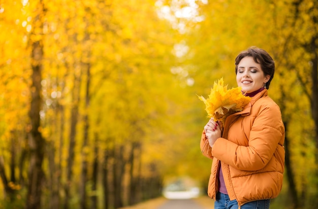 Cheerful young girl with short brown hair wearing autumn yellow jacket walking at the park Autumn walk Woman portrait Girl is playing with leaf and smiling Woman enjoying fall nature