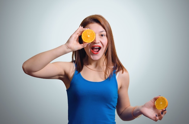 Cheerful young girl with a slice of orange around the eyes on the gray studio wall.