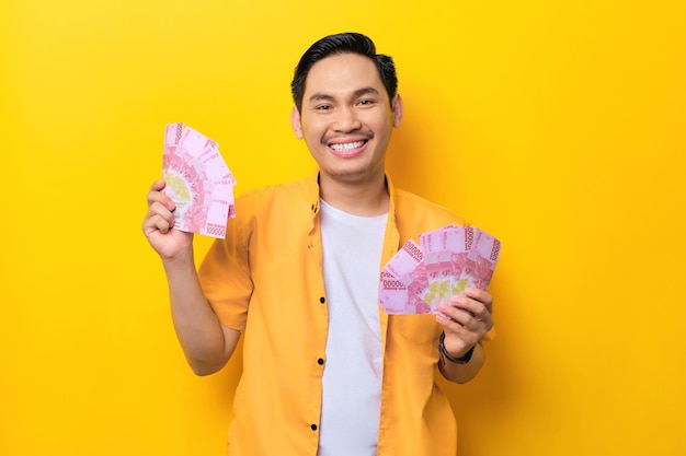 Cheerful young handsome Asian man holding bunch of money banknotes isolated on yellow background