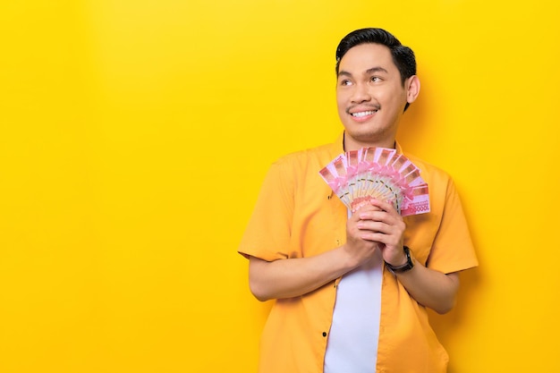 Cheerful young handsome Asian man holding bunch of money and looking away at copy space isolated on yellow background