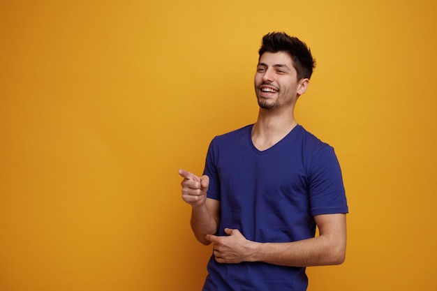 Cheerful young handsome man looking and pointing at side laughing on yellow background with copy space