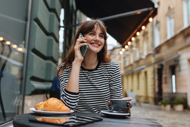 Cheerful young lady in casual clothes enjoying a fresh coffee and talking on mobile phone