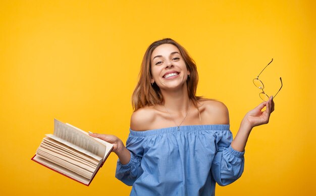 Photo cheerful young lady with book and eyeglasses smiling at camera in yellow studio