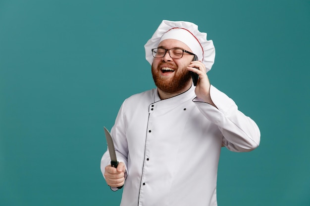 Cheerful young male chef wearing glasses uniform and cap holding knife laughing with closed eyes while talking on phone isolated on blue background