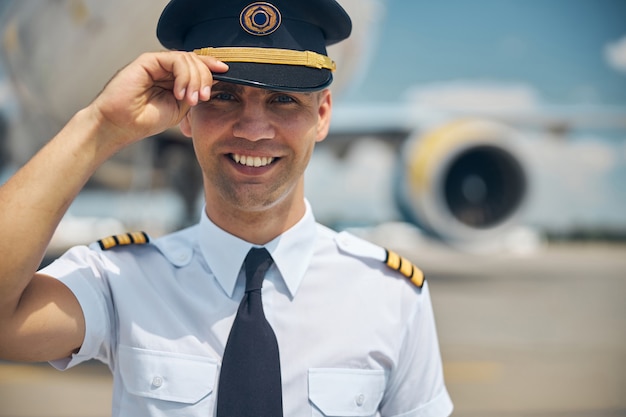 Cheerful young man airline worker in pilot cap standing outdoors at airport