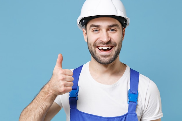 Cheerful young man in coveralls protective helmet hardhat isolated on blue wall  portrait. Instruments accessories for renovation apartment room. Repair home concept. Showing thumb up.