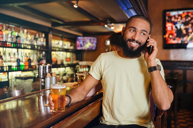 Cheerful young man drinking beer and talking on mobile phone in pub