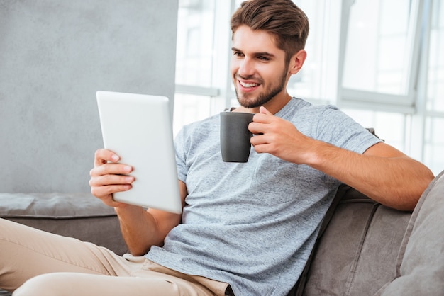 Cheerful young man in grey t-shirt sitting on sofa at home. Communication by tablet and smiling while drinking a coffee.