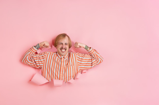 Cheerful young man poses in torn coral paper hole wall emotional and expressive