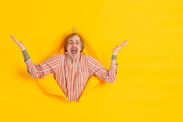 Photo cheerful young man poses in torn yellow paper hole wall emotional and expressive