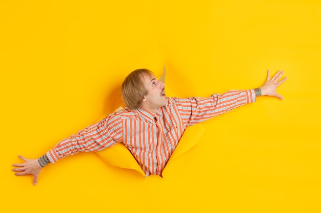 Cheerful young man poses in torn yellow paper hole wall emotional and expressive