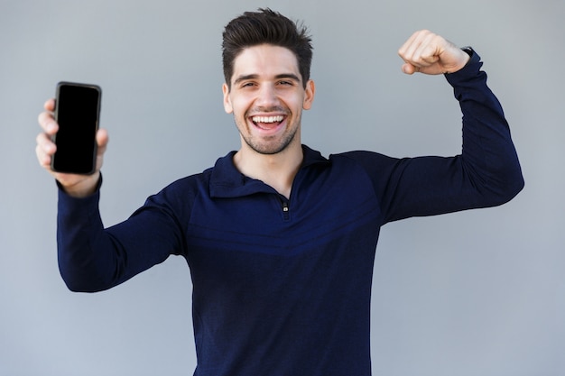 Cheerful young man showing blank screen mobile phone while standing isolated over gray