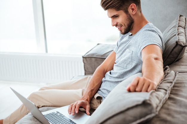 Cheerful young man in t-shirt sitting on sofa at home. Working on laptop computer and smiling.