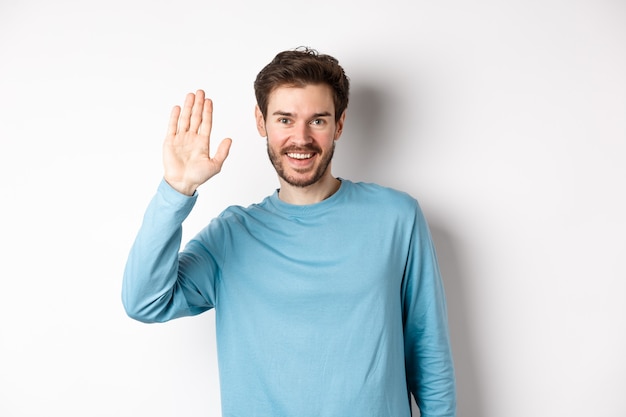 Cheerful young man with beard saying hello, looking friendly and waving hand to greet you, standing over white background